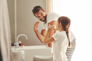 Dental care, father with daughter brush their teeth and in bathroom of their home. Oral hygiene routine, parent with child use toothbrush for health and wellness mouth protection in the morning.